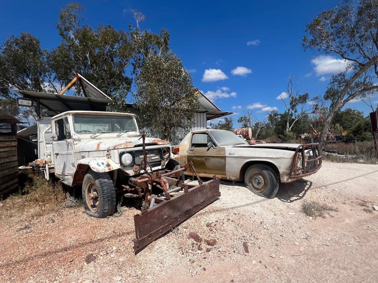 Two weathered vehicles on an opal field property: a rusted Toyota Land Cruiser with a plough blade on the front, and an old sedan with a bull bar