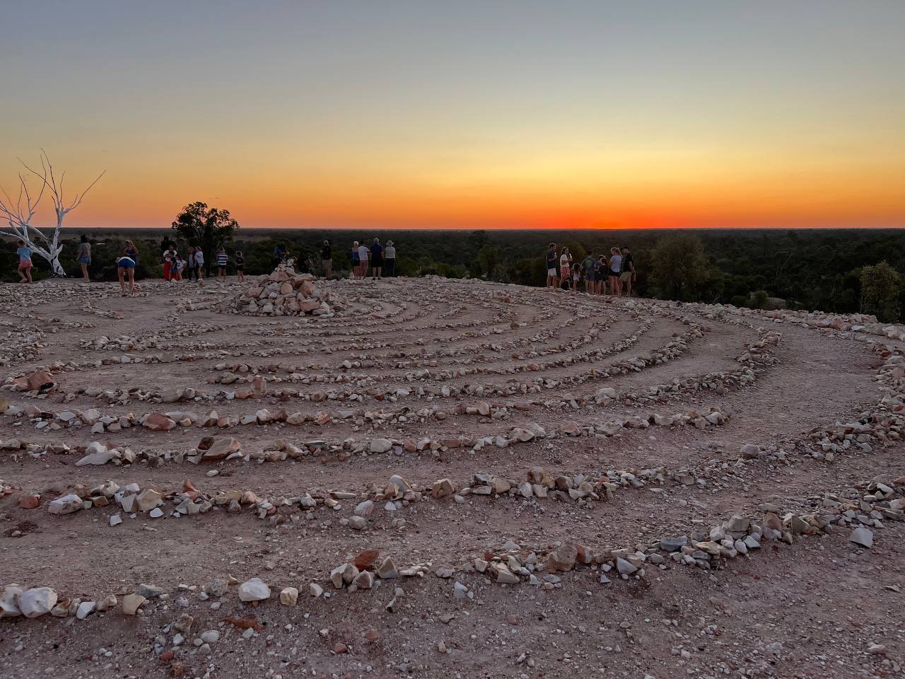 A large circular stone labyrinth on the hilltop, with people walking its paths as the sun sets