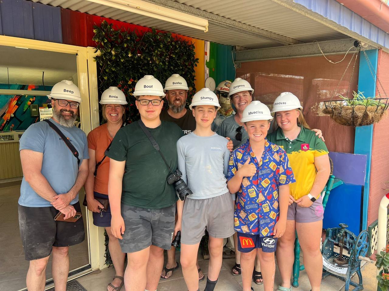 The full touring party of nine in white hard hats outside the mine, the hats labelled Beep, Rosie, Boof, Buster, Stumpy, Gorgeous, Muscles, and Princess